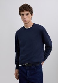 Mörkblå stickad tröja med rund halsringning, långa ärmar och en avslappnad passform, bärs över en vit skjorta, kombinerad med mörka jeans.