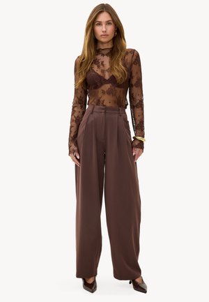 Woman wearing sheer brown lace long-sleeve top, high-waisted wide-leg brown trousers, and pointed brown heels, standing against a plain background.