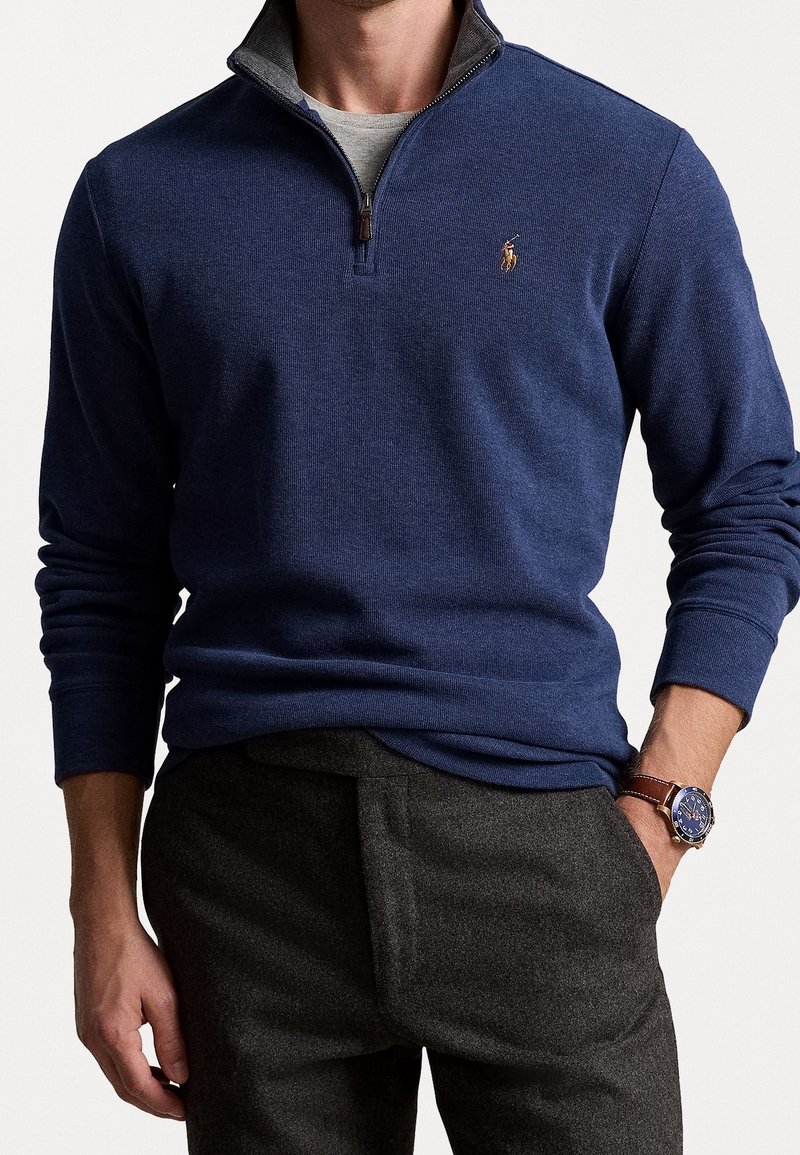 Homme portant un pull à fermeture éclair quart bleu marine avec logo brodé, chemise grise en dessous, pantalon foncé et montre avec bracelet en cuir marron.