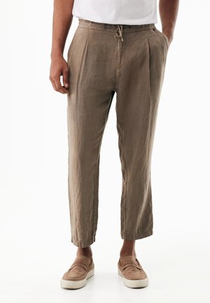 Hombre con pantalones casuales beige con cordón y zapatos de ante a juego sin cordones, de pie con una mano en el bolsillo.