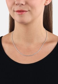 Närbild av en kvinna som bär ett tunt silverhalsband och en svart topp, med läppar som ler svagt, rakt hår som är bakåttrollat.