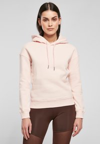 Rosa hettegenser med snorhette, ribbestrikkede mansjetter og kant, laget av mykt materiale. Båret med brune leggings, som viser en avslappet passform.