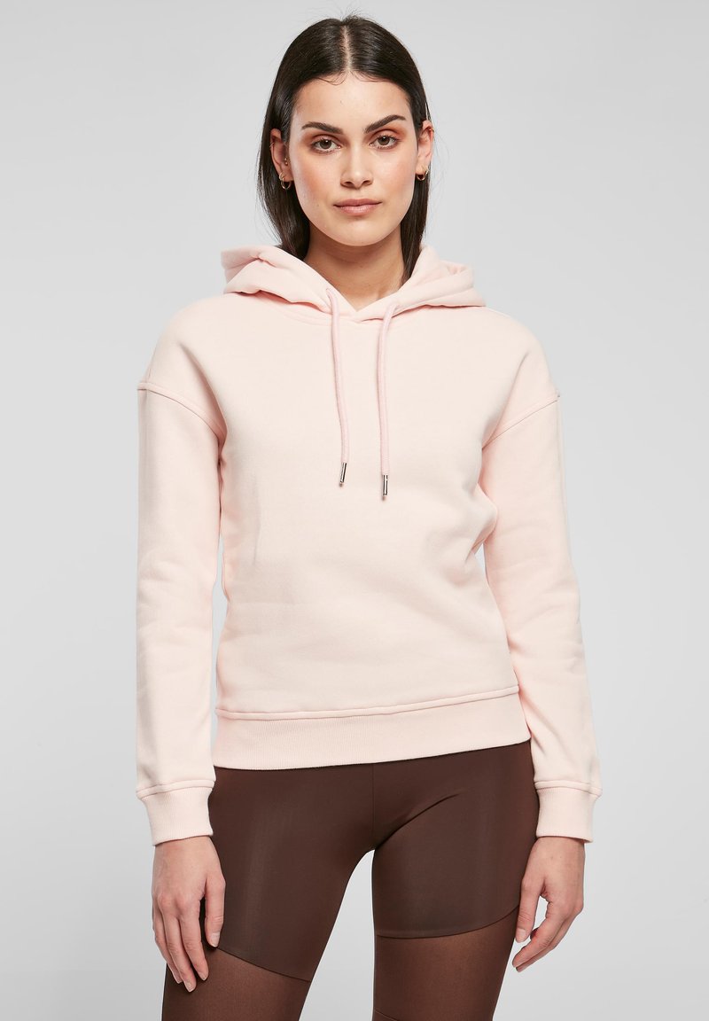 Rosa hettegenser med snorhette, ribbestrikkede mansjetter og kant, laget av mykt materiale. Båret med brune leggings, som viser en avslappet passform.