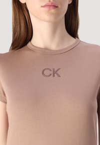 Šviesiai rudos spalvos trumpomis rankovėmis marškinėliai iš minkštos medžiagos, su įrūdytu spalvotu kaklu ir siuvinėtu "CK" logotipu ant krūtinės.
