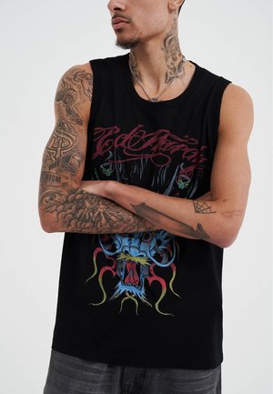 Modèle masculin avec des tatouages, portant un débardeur noir à manches courtes présentant un artwork coloré de dragon, bras croisés, fond neutre.