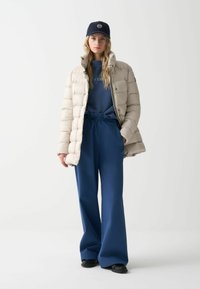 Chaqueta acolchada beige con acabado brillante, sudadera azul y pantalones azules holgados. El conjunto se completa con una gorra oscura y calzado negro.