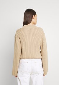 Beżowy sweter dzianinowy z żebrowaną fakturą, długimi rękawami i wysokim dekoltem. Noszony z białymi spodniami, widziany od tyłu.