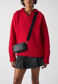 Sarkans adīts džemperis ar apkakles izgriezumu un ribinātu apakšmalu, saskaņots ar melnu patterned plecu somu un svītrainu mini svārku.