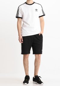 Man wearing Adidas white T-shirt with black stripes, black shorts, and black sneakers standing with one hand in pocket.