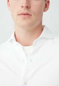 Witte overhemdd met kraag, met een gladde textuur en subtiele details bij de knopen. Gelaagd over een witte ronde hals.