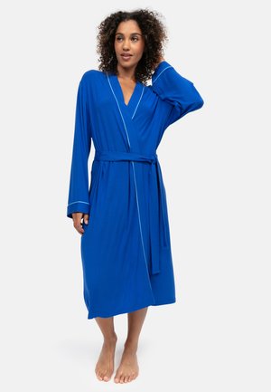 Femme aux cheveux bouclés debout pieds nus, portant une longue robe bleue avec des bordures blanches et une ceinture à la taille, sur un fond uni.