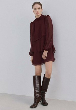 Mujer con vestido burdeos de manga larga de capas y botas de cuero marrón oscuro hasta la rodilla, de pie contra una pared lisa y clara.