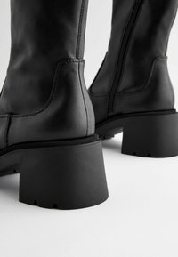 Botas de tobillo de cuero negro con un tacón grueso y texturizado. Presentan un cierre de cremallera, costuras sutiles y un acabado suave.