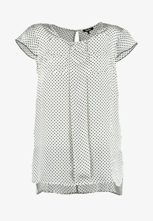 Vrouw lopend, gekleed in een witte blouse met polkadots, zwarte broek, witte schoenen, en een kleine witte clutch vasthoudend tegen een effen achtergrond.