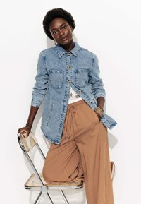 Femme appuyée sur une chaise, portant une veste en denim bleu, un haut en dentelle blanche et un pantalon large rayé marron, avec une main dans la poche.