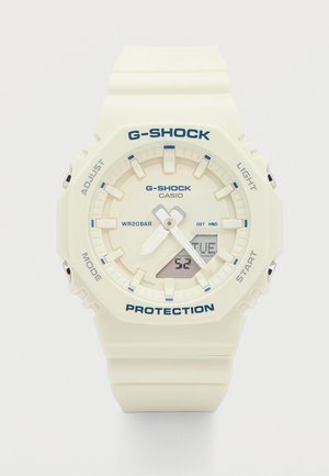 Valge Casio G-Shock käekell analoogosutitega, digitaalne kuupäeva näidik, mis kuvab "TUE", ja kaitsev vahtkummist rihm.