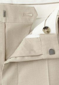 Ceinture de pantalon beige avec bouton, fermeture éclair, fermeture crochet et barre, et doublure intérieure blanche partiellement visible.