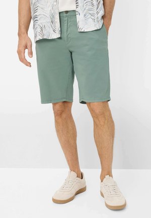 Man draagt lichtgroene knielange short, witte sneakers en een kortemouwen overhemd met een bladmotief.