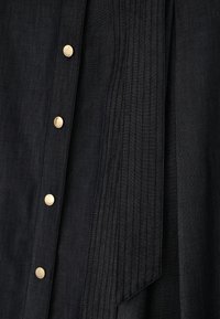 Zbliżenie czarnego materiału z pionowymi zakładkami i rzędem czterech błyszczących złotych guzików wzdłuż krawędzi.