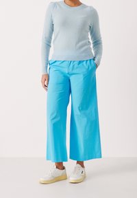 Persoon draagt lichtblauwe trui, felblauwe wijd uitlopende broek, witte sneakers, staat met handen in zakken op een witte achtergrond.