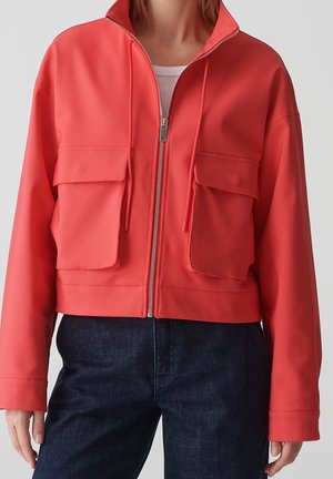 Femme portant une veste corail zippée avec deux grandes poches à rabat à l'avant, associée à un jean bleu foncé et un top blanc en dessous.
