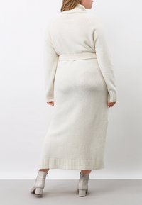 Lång, krämfärgad stickad kappa med hög krage, figursydd midjesnodd, ribbade ärmslut och nederkant. Mjuk textur och avslappnad silhuett.