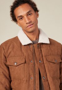 Brun mockaläderjacka med en vit shearlingkrage, dragkedja fram och två bröstfickor med silverdetaljer. Mjuk textur och kviltad design.