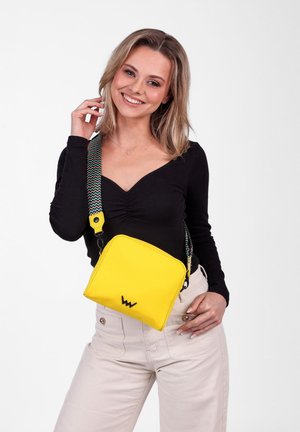 Yellow square crossbody bag made of durable fabric, featuring a patterned strap, logo detail, and a zip closure.