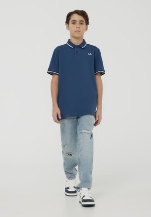 Granatowa koszulka polo z białymi akcentami, krótkie rękawy i zapięcie na trzy guziki, w połączeniu z jasnoniebieskimi, zniszczonymi dżinsami i trampkami.