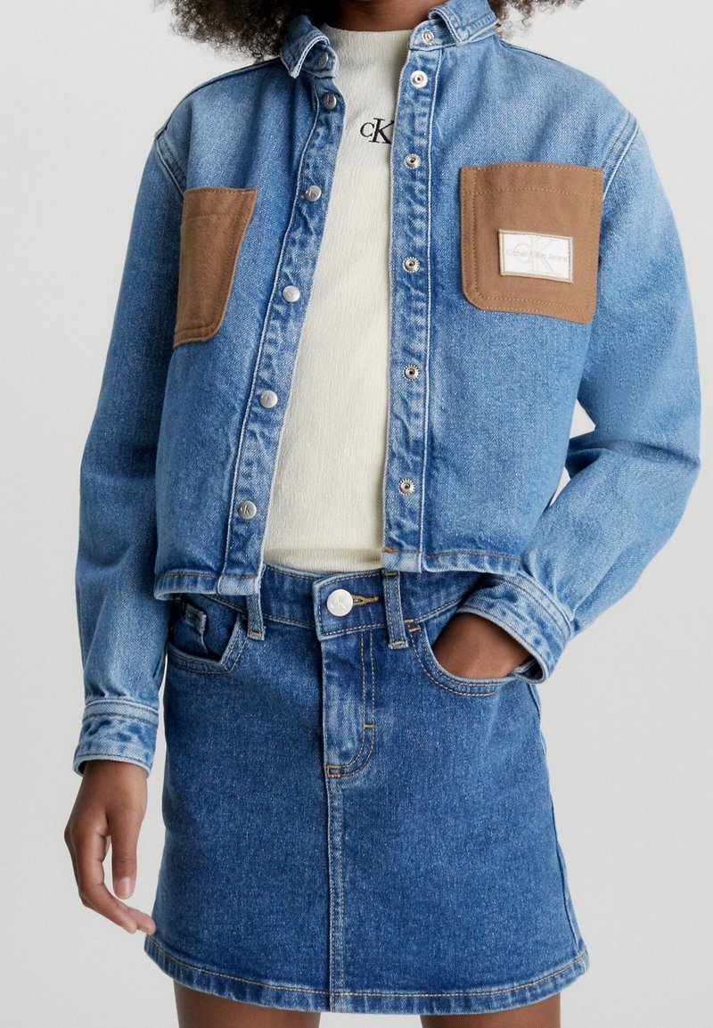 Person som bär en blå denimjacka med bruna fickor över en vit Calvin Klein-tröja och en matchande blå denimkjol, med handen i fickan.