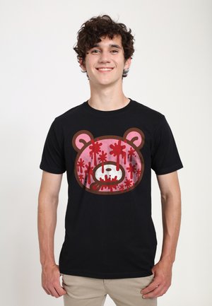Ung mand iført sort t-shirt med en pink bamseansigt, der er sprøjtet med rød maling, smilende med hænderne i siden.