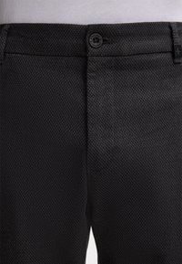 Black textured trousers featuring a subtle diamond pattern, a button closure, and side pockets, made from a smooth fabric.