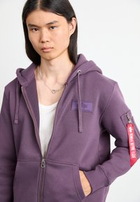 Felpa con zip di colore viola con una finitura testurizzata, tasche frontali, cappuccio regolabile e un'etichetta di tessuto rossa con la scritta "RIMUOVERE PRIMA DEL VOLO" sulla manica.
