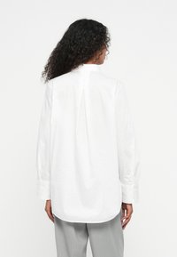 Biała koszula z długim rękawem z plisą na plecach, subtelnym wzorem kropek i zapięciem na guziki przy mankietach, w zestawieniu z jasnymi spodniami w paski.