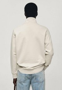 Pulover bej deschis cu guler înalt, manșete și tiv cu model țesut. Textură moale, design minimalist, purtat cu blugi albaștri deschiși.