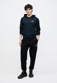 Sudadera con capucha azul marino con bolsillo delantero, capucha con cordón y logo. Combinada con pantalones jogger negros con un logo sutil, ambos de tela suave. Zapatillas negras.