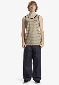 Top de tirantes a rayas en tono marrón con líneas azules, ribete negro y logotipo; combinado con jeans de corte holgado en azul marino y zapatillas negras.