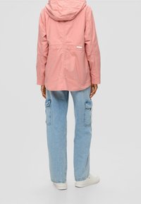 Osoba oblečená v světle růžové bundě s kapucí a světle modrých džínových kalhotách s kapsami, stojící zády k přední straně na jednobarevném pozadí.