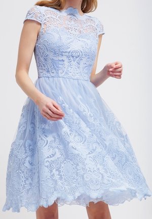 Vrouw met een lichtblauwe kanten jurk met korte mouwen, een aansluitend lijfje en een uitlopende rok met ingewikkeld bloemrijk borduurwerk.