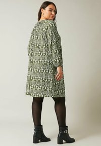 Grünes und weißes gemustertes Kleid mit langen Ärmeln, knielanger Schnitt, strukturierter Stoff, kombiniert mit schwarzen Knöchelstiefeln.