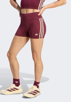 Osoba ubrana w bordowe spodenki Adidas, dopasowany krótki top, bordowe skarpetki i białe buty Adidas na jasnym tle.