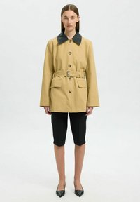 Cappotto beige con cintura, colletto nero, grandi bottoni e due tasche frontali, abbinato a pantaloni corti neri e scarpe col tacco nere a punta.