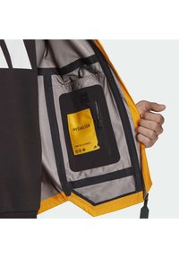 Main tenant une veste jaune ouverte montrant une poche intérieure avec une fermeture éclair noire et une étiquette jaune portant l'inscription "MY SHELTER" et "OWN THE ELEMENTS".