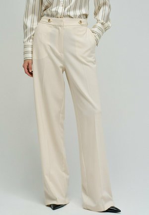 Mujer con pantalones anchos de tiro alto color crema con botones frontales y una blusa a rayas metida, de pie con una mano en el bolsillo.