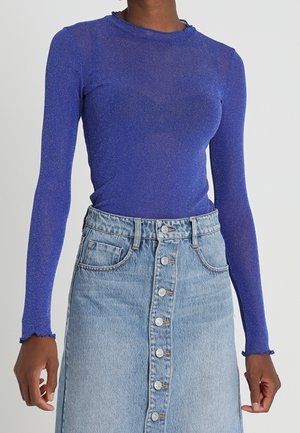 Femme portant un haut bleu royal transparent à manches longues rentrée dans une jupe en denim clair taille haute à boutons sur le devant.