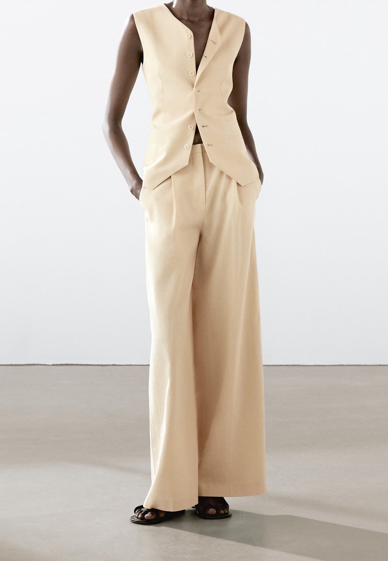 Modèle portant un gilet beige sans manches boutonné et un pantalon large assorti, les mains dans les poches, debout sur un sol gris devant un mur blanc.