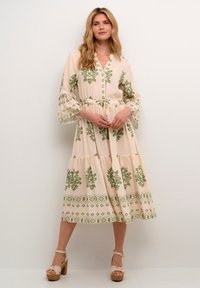 Long, cream dress with green floral patterns, gathered waist, button front, and flared sleeves. Worn with wedge heels.