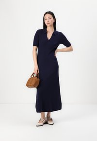 Sweater dress in navy blue, ribbed fabric, short sleeves, V-neck, floor-length. Holding a small brown, textured handbag. Beige shoes with accents.