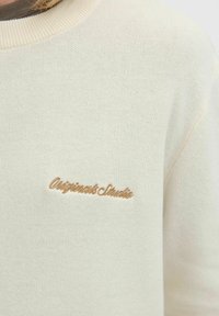 Helle beigefarbene Sweatshirt mit geripptem Rundhalsausschnitt. Mit gesticktem "Originals Studio"-Logo in goldener Fäden auf der linken Brust. Weiche Textur.