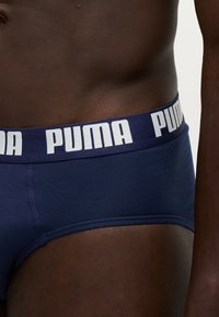 Κοντινό πλάνο ενός ατόμου που φοράει σλιπ Puma σε σκούρο μπλε με σκούρο μπλε ζώνη μέσης και λευκό κείμενο λογότυπου Puma.